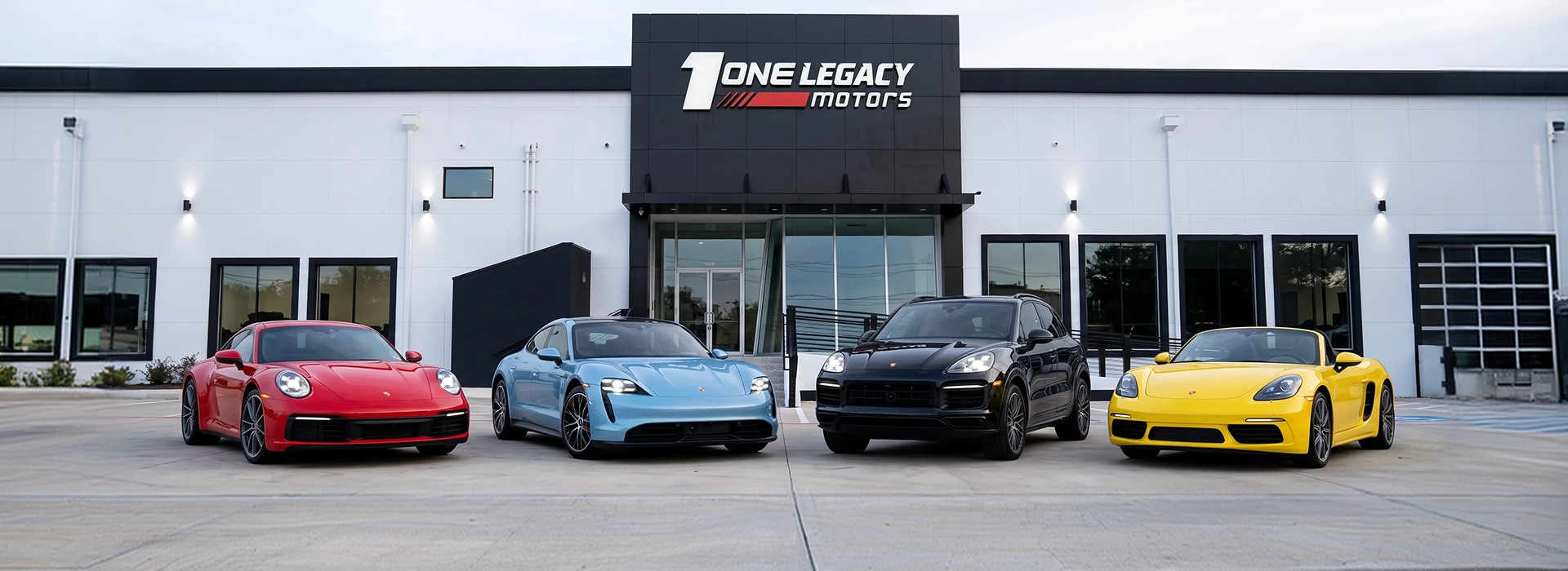 A red Porsche 911, a blue Taycan, a black Cayenne and a yellow 718 Boxster parked in a row outside the One Legacy Motors showroom in Carrollton, Texas