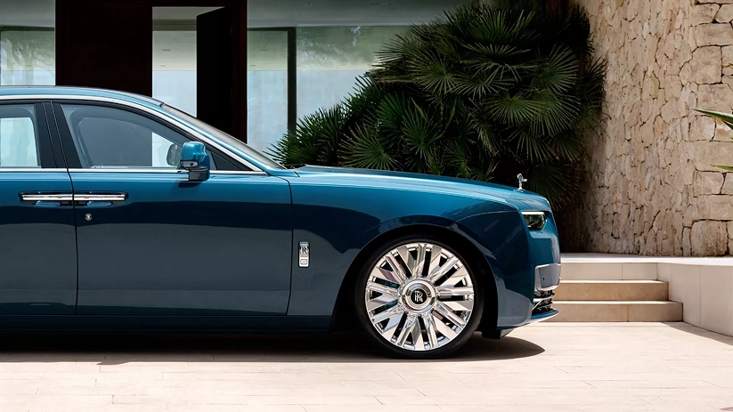 Two-tone blue Rolls-Royce Ghost in profile beside a stone wall and palms