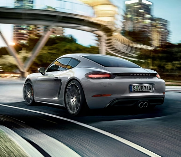 Silver Porsche 718 Cayman seen from the rear three-quarter at speed under a curved pedestrian bridge