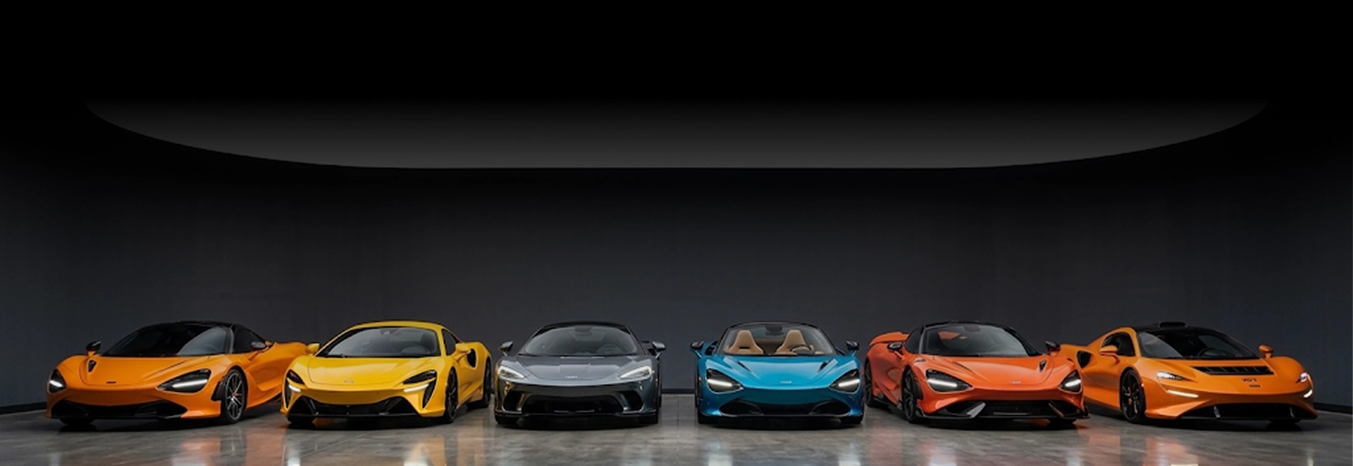 Six McLaren supercars in orange, yellow, grey, blue and orange lined up under a curved light in a dark studio