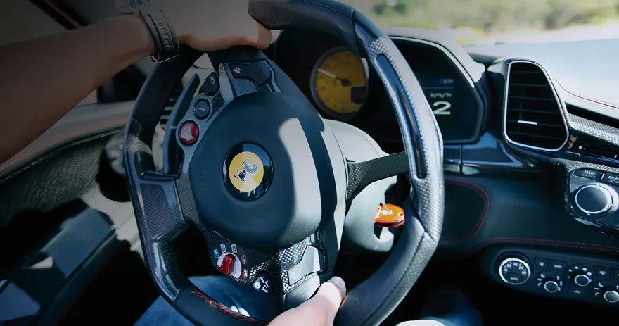 Driver's hand on the yellow-tachometer steering wheel of a Ferrari 458 Spider on a sunlit road